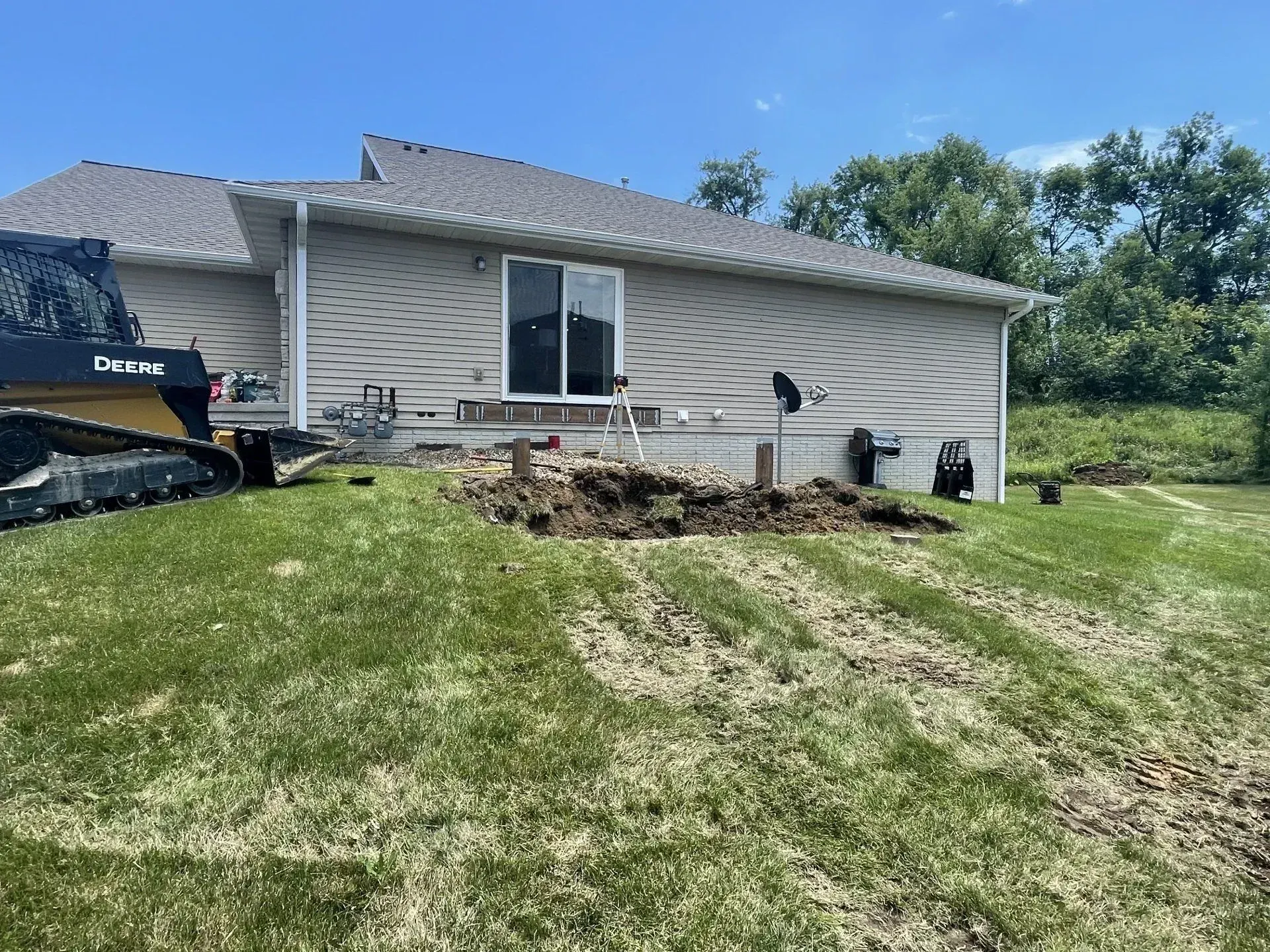 Compact track loader beside a freshly dug patch of soil in the backyard lawn of a gray ranch house.