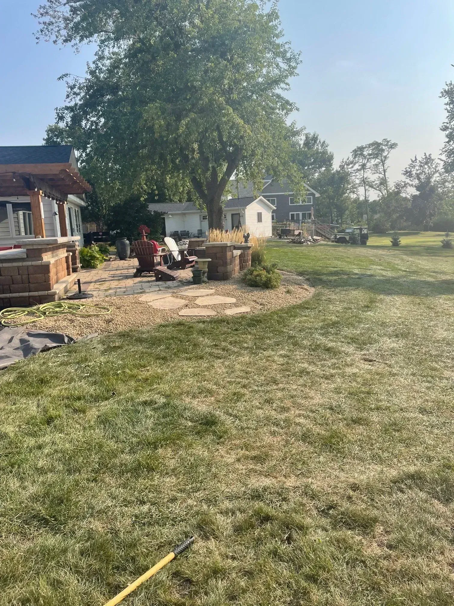 Backyard stone patio with a fire pit and pergola beside a large shade tree on a sunny day.