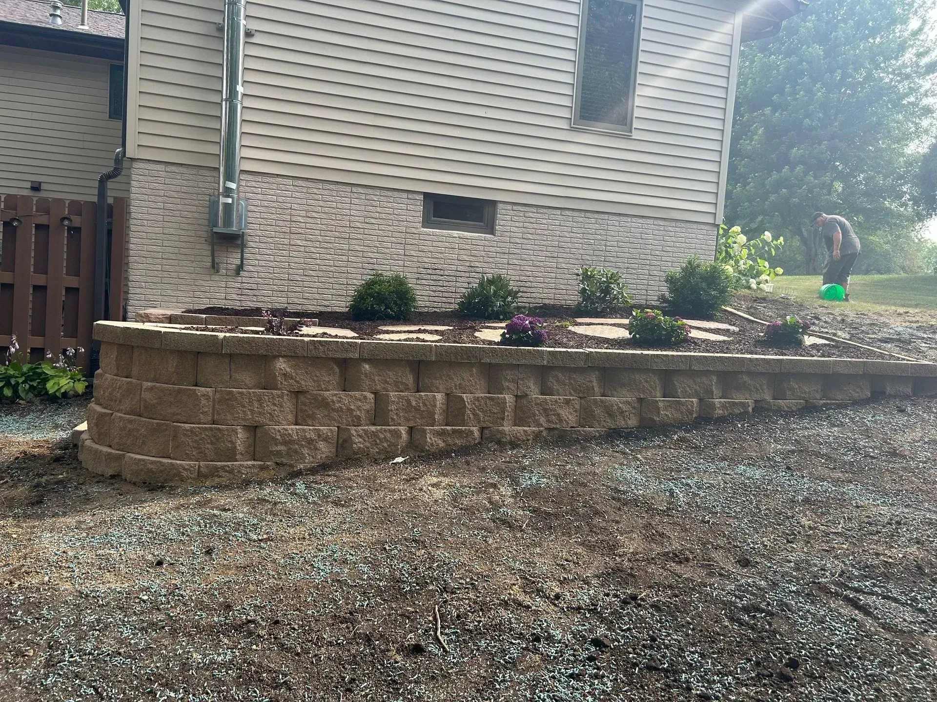 Retaining wall made of tan blocks holds a flower bed with bushes. House and gravel driveway in background.