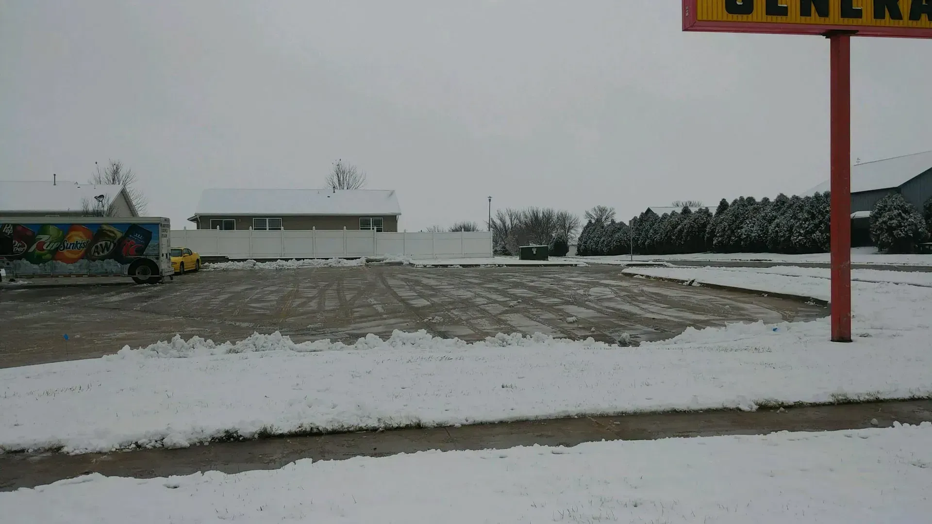 Plowed commercial parking lot with snow piled along the edges, a store sign on the right, and overcast skies.