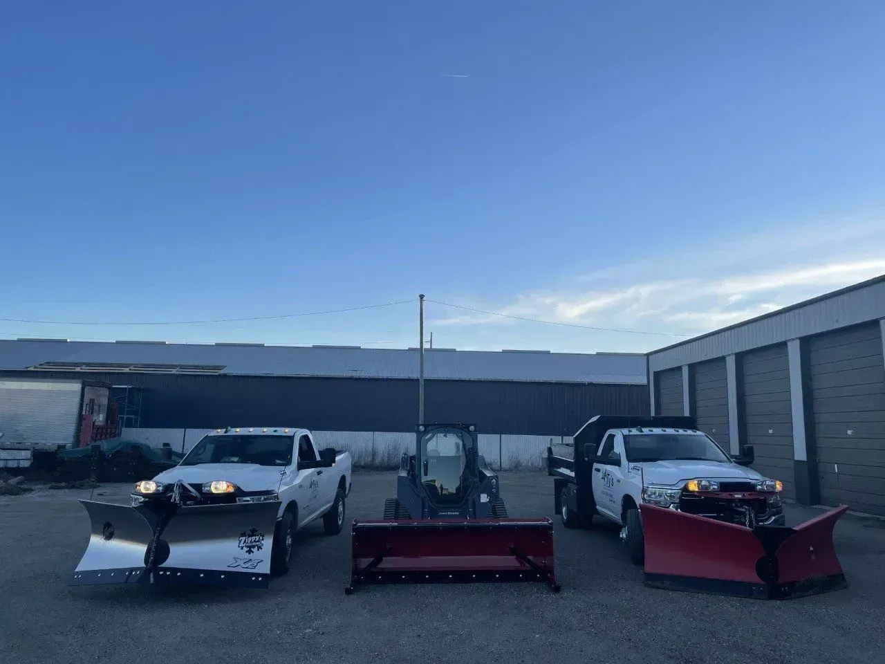 Two white pickup trucks and a skid steer fitted with snow plows, parked outside storage buildings.