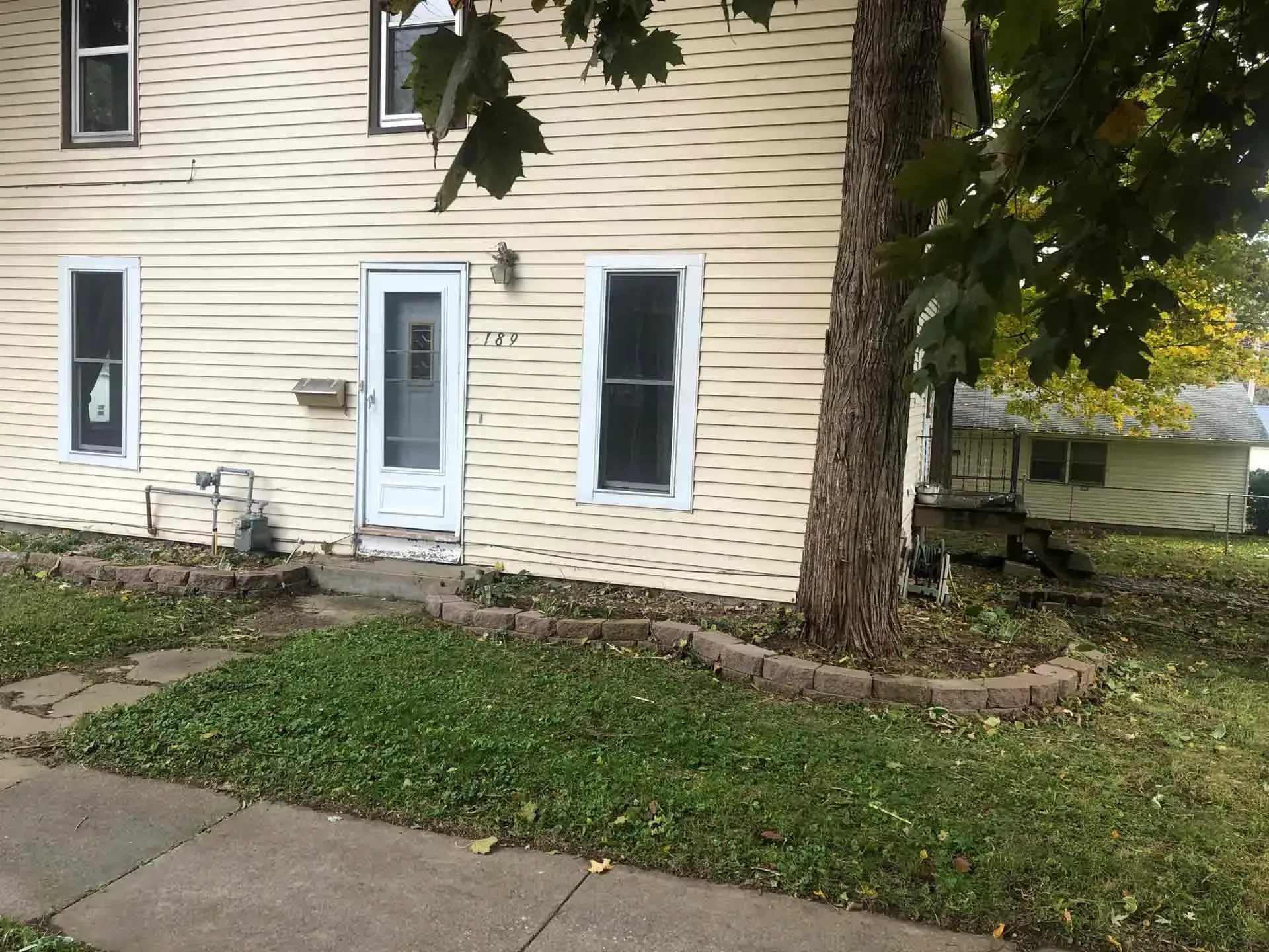 Two-story beige house with white trim and a front yard with a brick border around a tree.